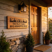the front entrance of a wooden house with a sign that reads "The Riley Family 8012 Asshopper Drive". The house has a stone wall and a wooden door, and there are two potted plants on either side of the door.
