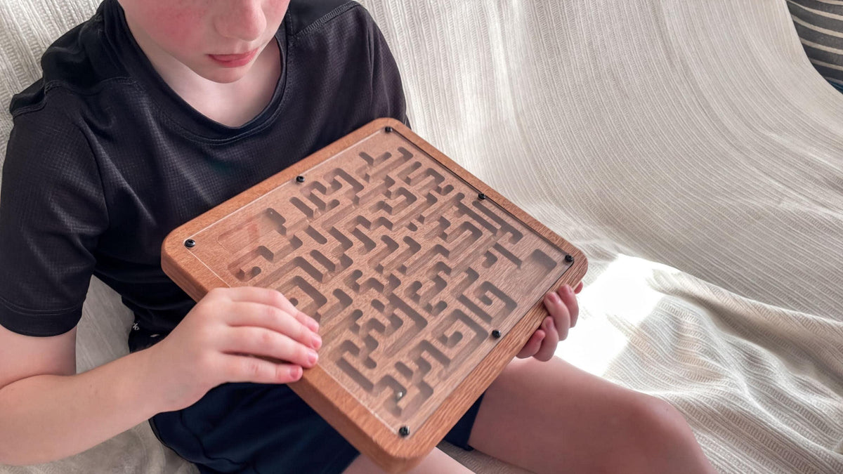 A young boy is sitting on a couch, holding a wooden maze game in his hands.