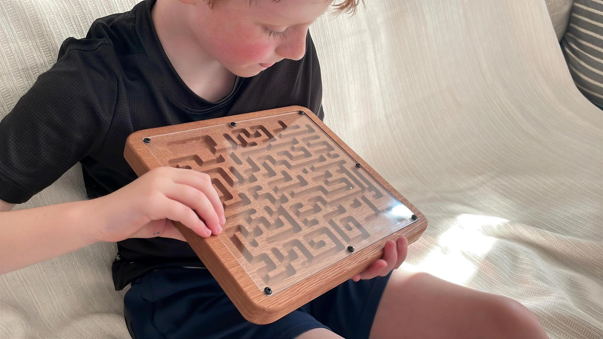A young boy is sitting on a couch, holding a wooden maze game board with a maze pattern on it.