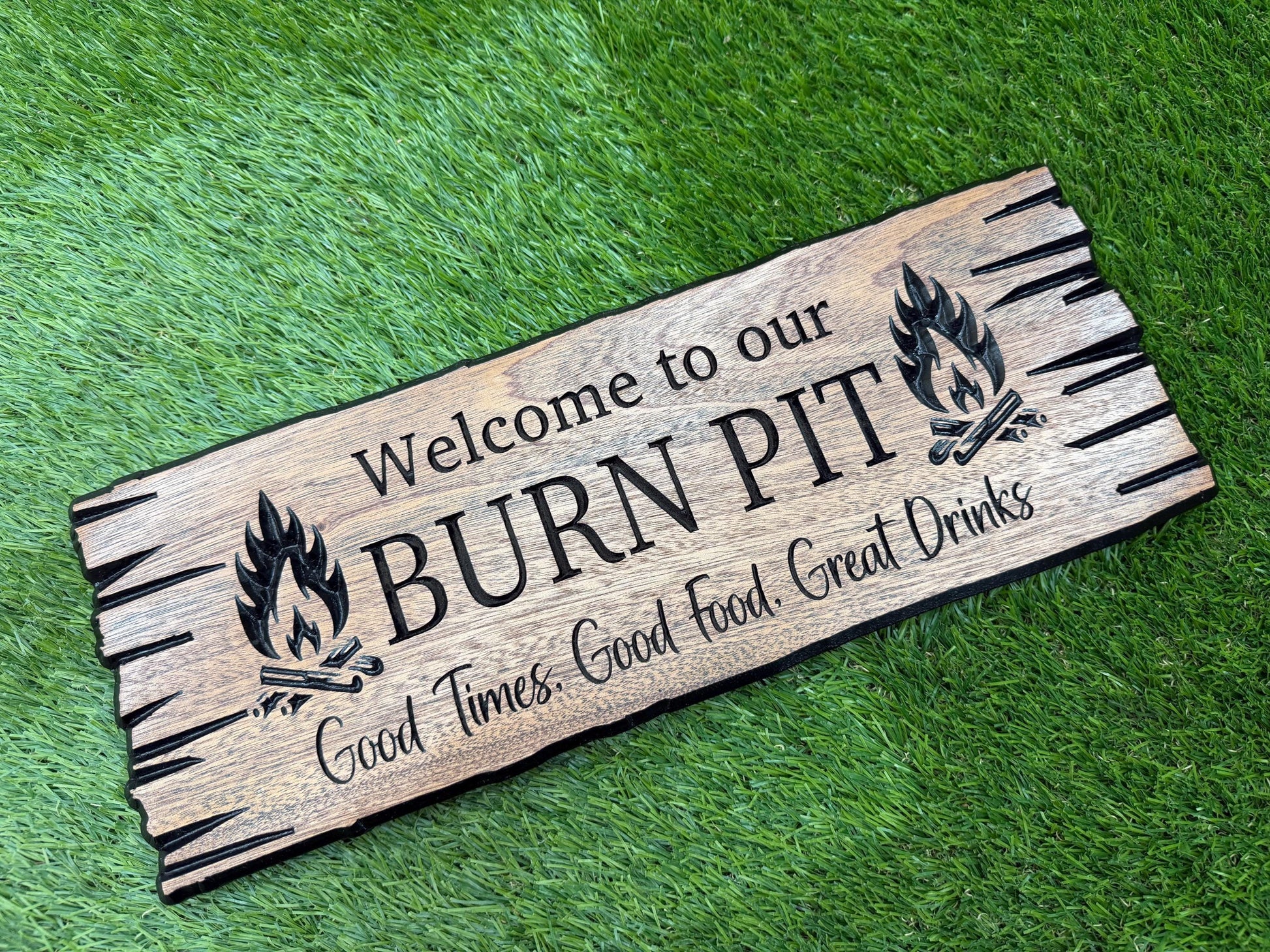 A wooden sign with the text "Welcome to our BURN PIT" and a graphic of a fire pit, placed on a grassy surface.