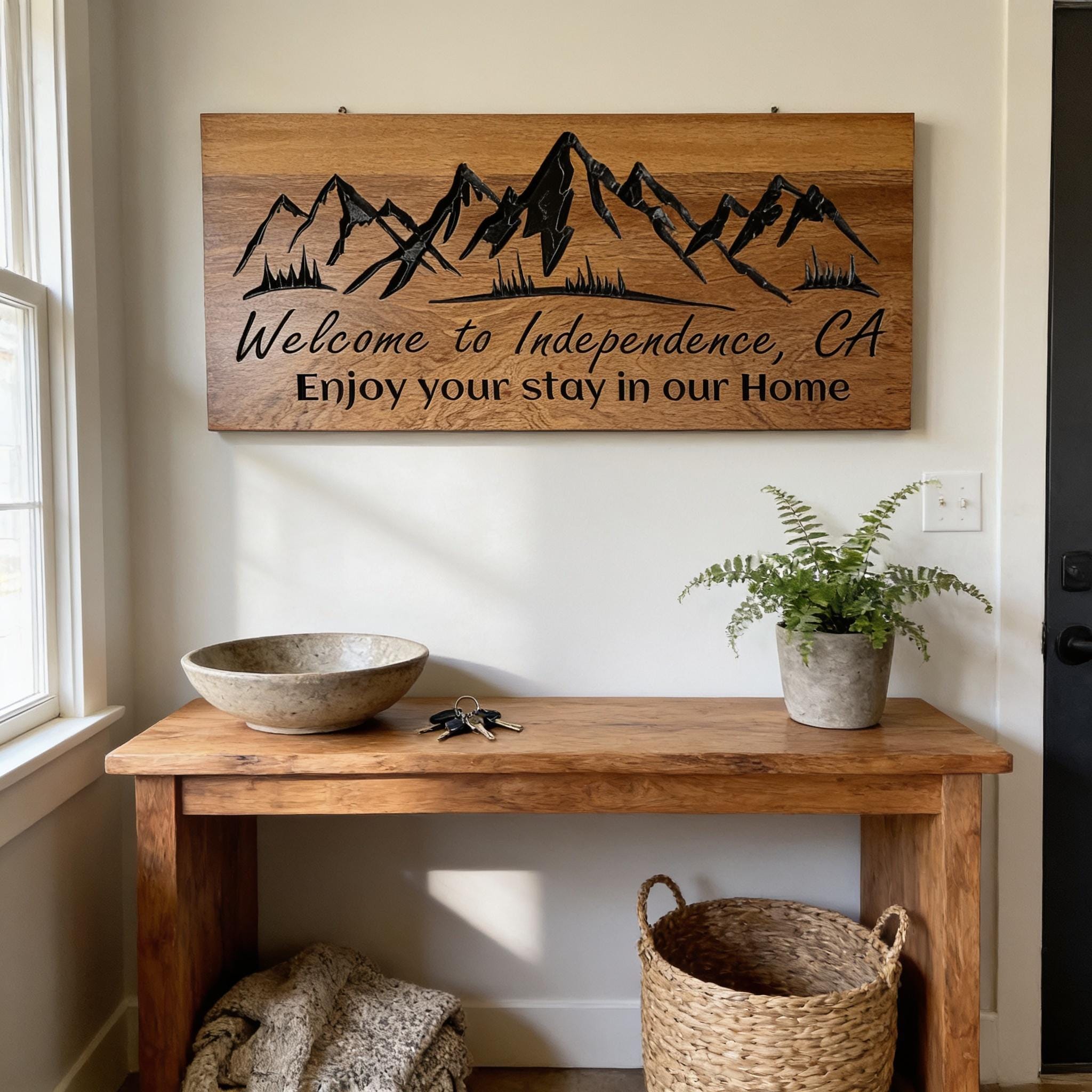 a wooden table with a bowl, a plant, and a woven basket, along with a wooden sign that reads "Welcome to Independence, CA Enjoy your stay in our Home".