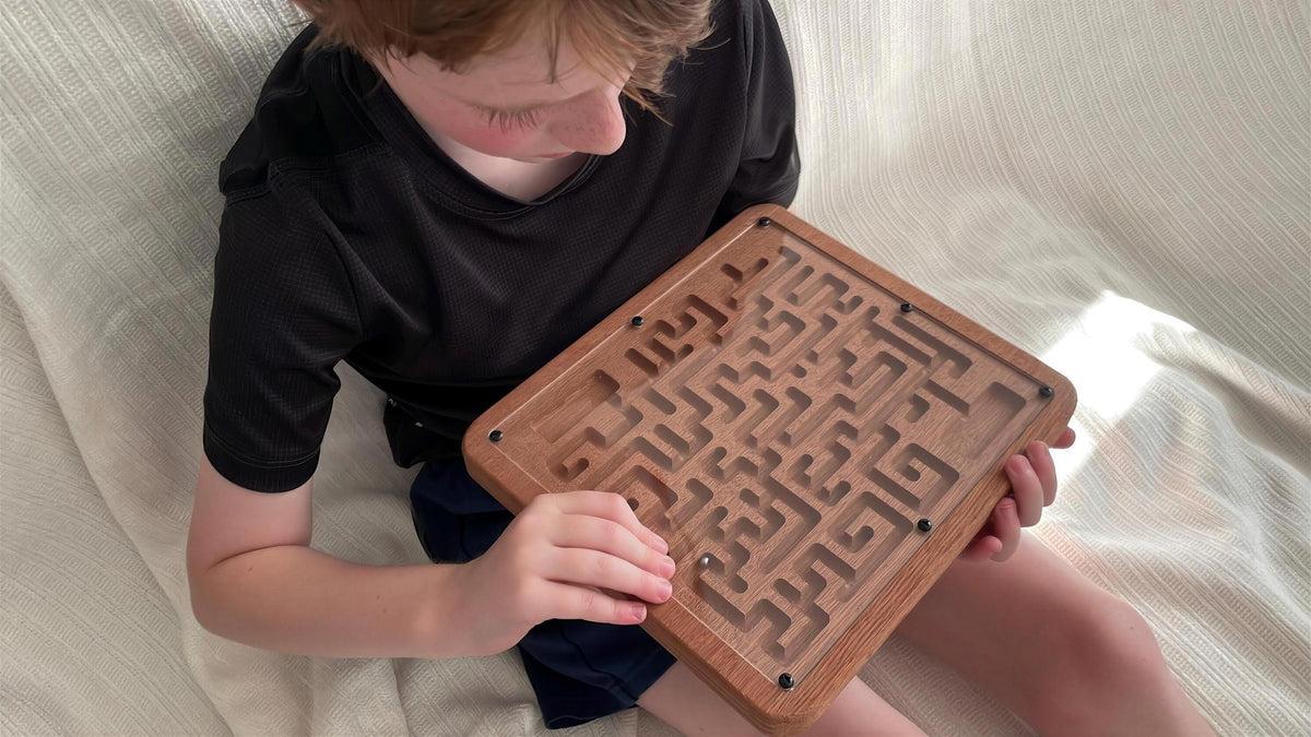 A young boy is sitting on a bed, holding a wooden maze puzzle in his hands.