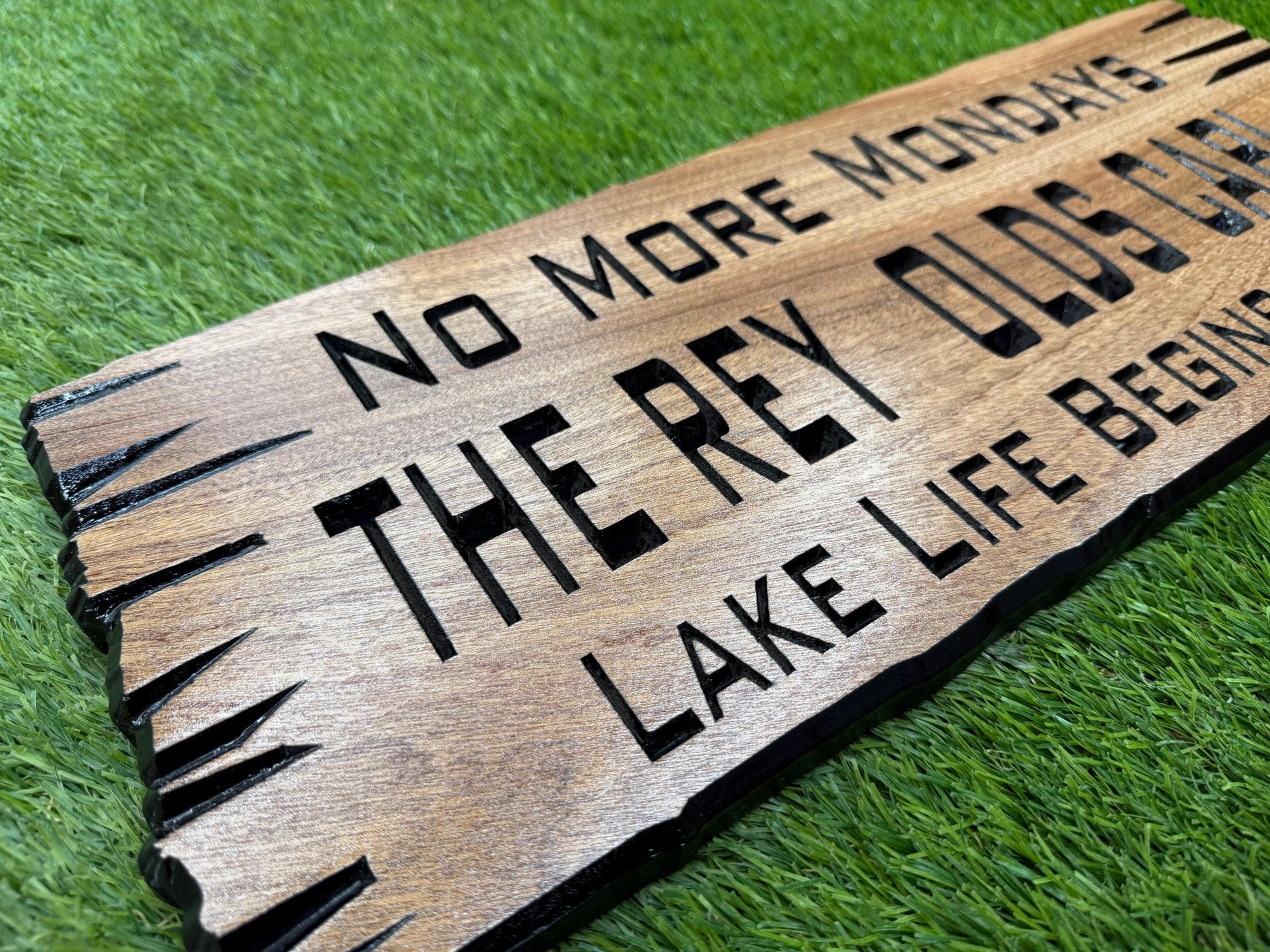 a wooden sign with the text "NO MORE MONDAYS" and "THE REY OLDS" carved into it, placed on a grassy surface.