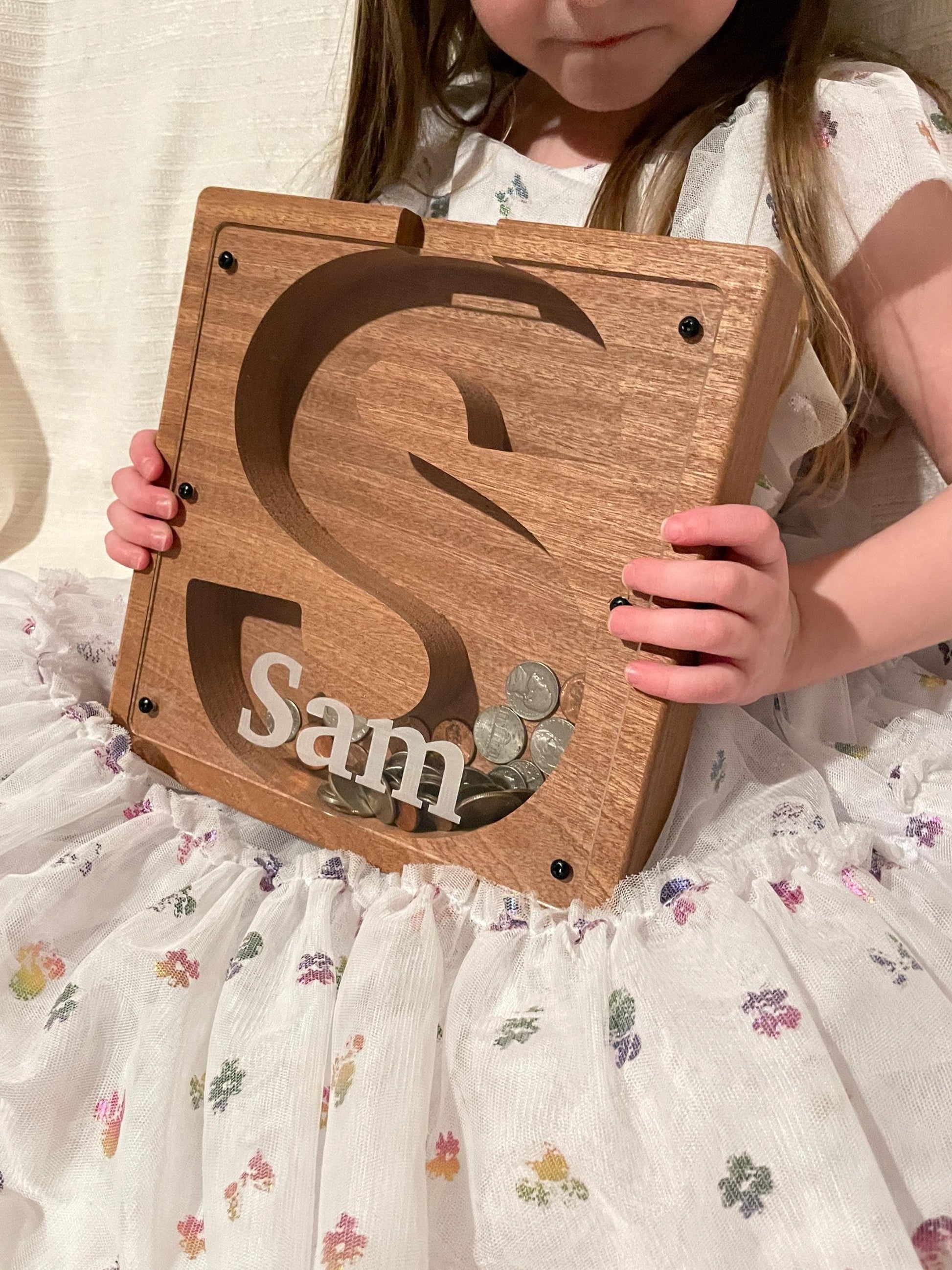 A young girl is holding a wooden coin box with the name "Sam" written on it.