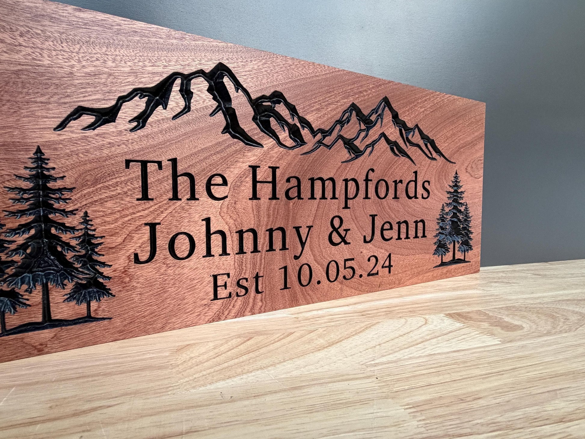 A wooden sign with a mountain landscape and trees, along with the names "The Hampfords" and "Johnny & Jenn" printed on it.