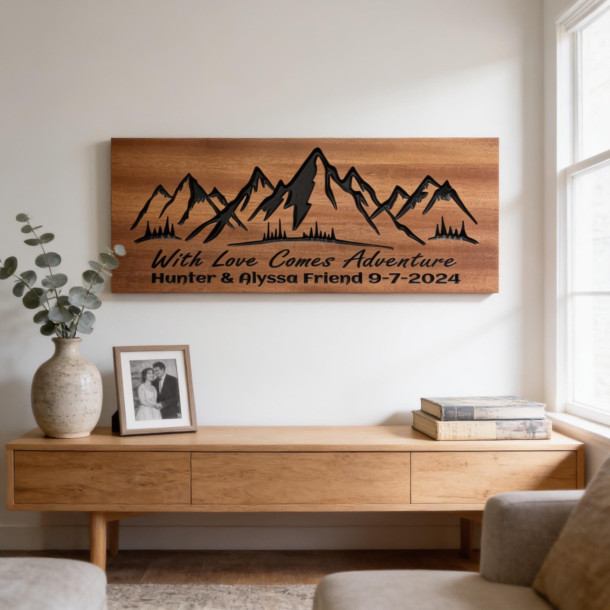 a wooden console table with a framed photo, a vase with greenery, and a wooden wall art depicting mountains. The wall art has the text "With Love Comes Adventure Hunter & Alyssa Friend 9-7-2024" written on it.