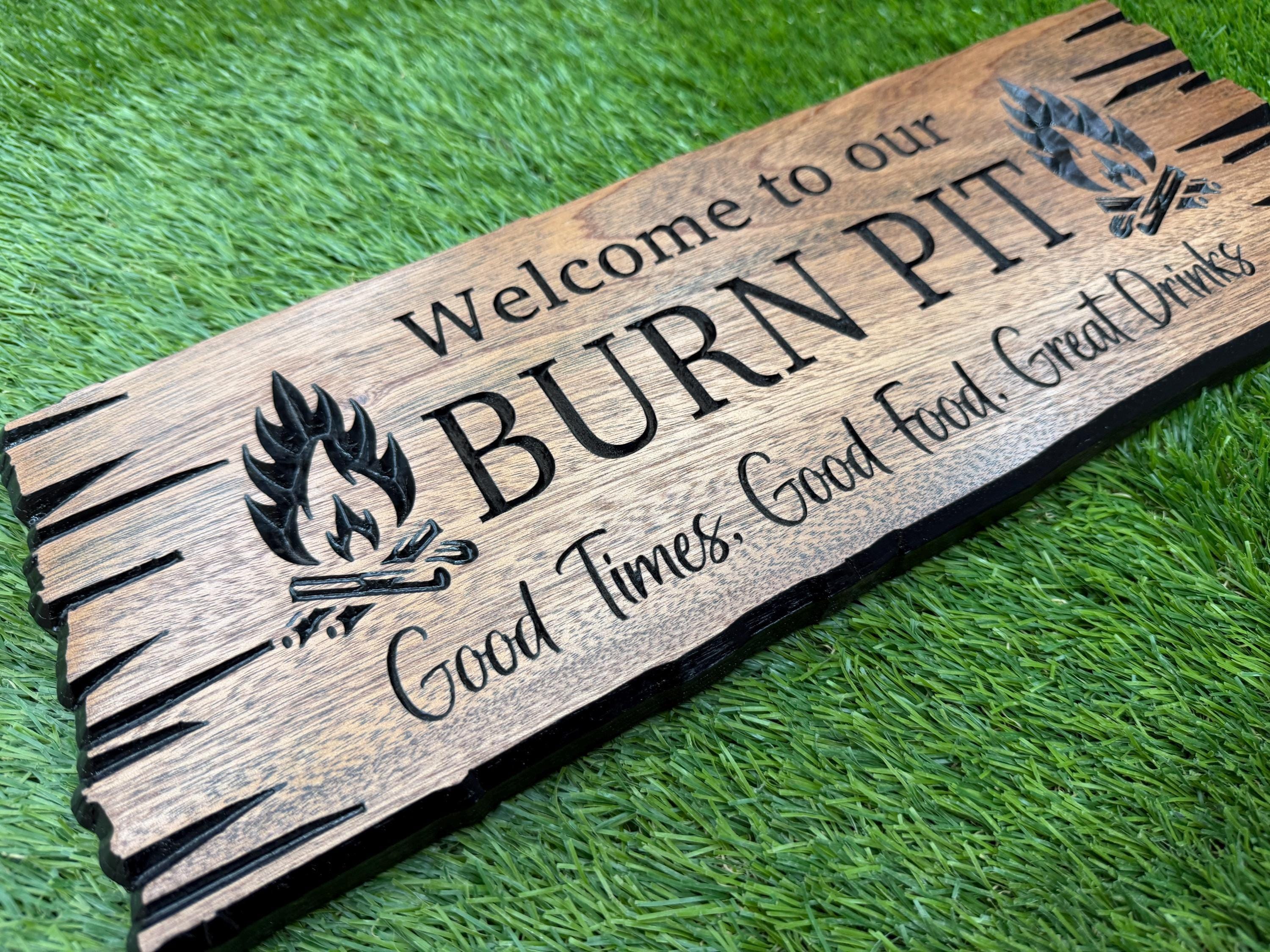 A wooden sign with the text "Welcome to our BURN PIT" and a graphic of a campfire, placed on a grassy surface.