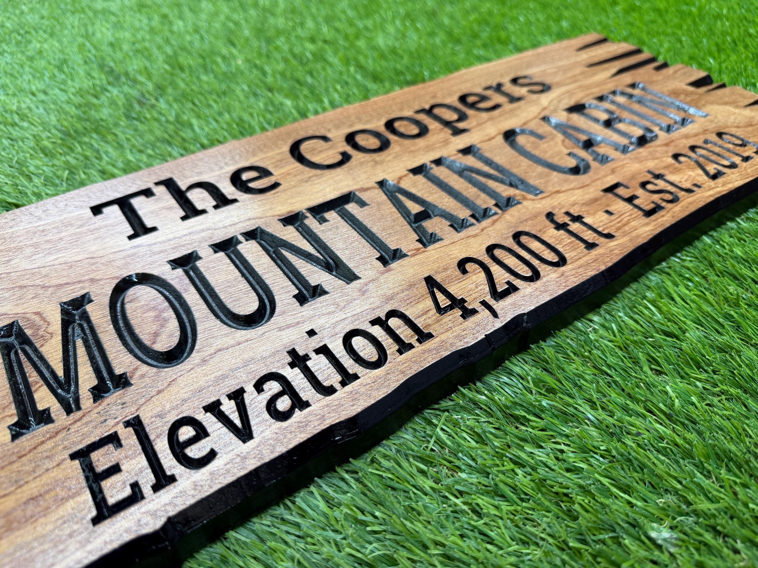 A wooden sign with the text "The Coopers Mountain Cabin Elevation 4,200 ft Est. 2019" engraved on it, placed on a grassy surface.
