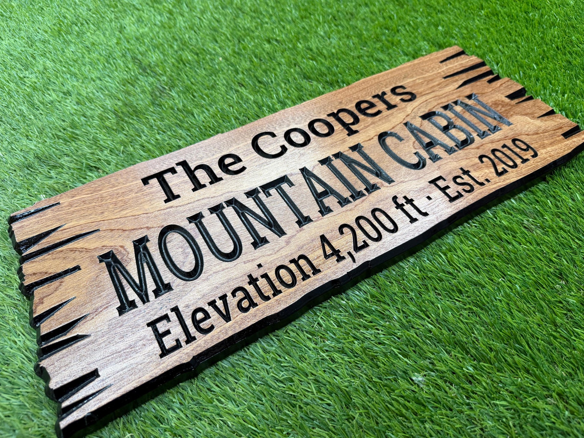 A wooden sign with the text "The Coopers Mountain Cabin Elevation 4,200 ft - Est. 2019" engraved on it, placed on a grassy surface.