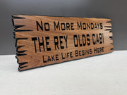 A wooden sign with the text "NO MORE MONDAYS THE REY OLDS CABI LAKE LIFE BEGINS HERE" carved into it.