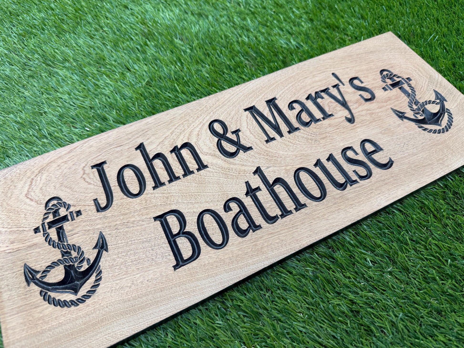 A wooden sign with the names 'John & Mary's Boathouse' engraved on it, placed on a grassy surface.