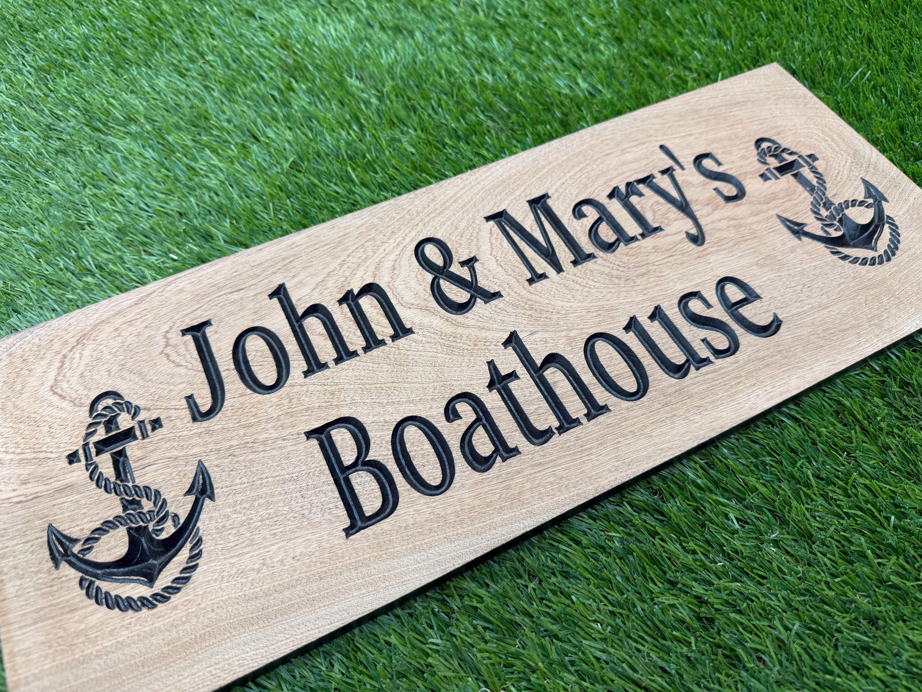A wooden sign with the names 'John & Mary's Boathouse' engraved on it, placed on a grassy surface.