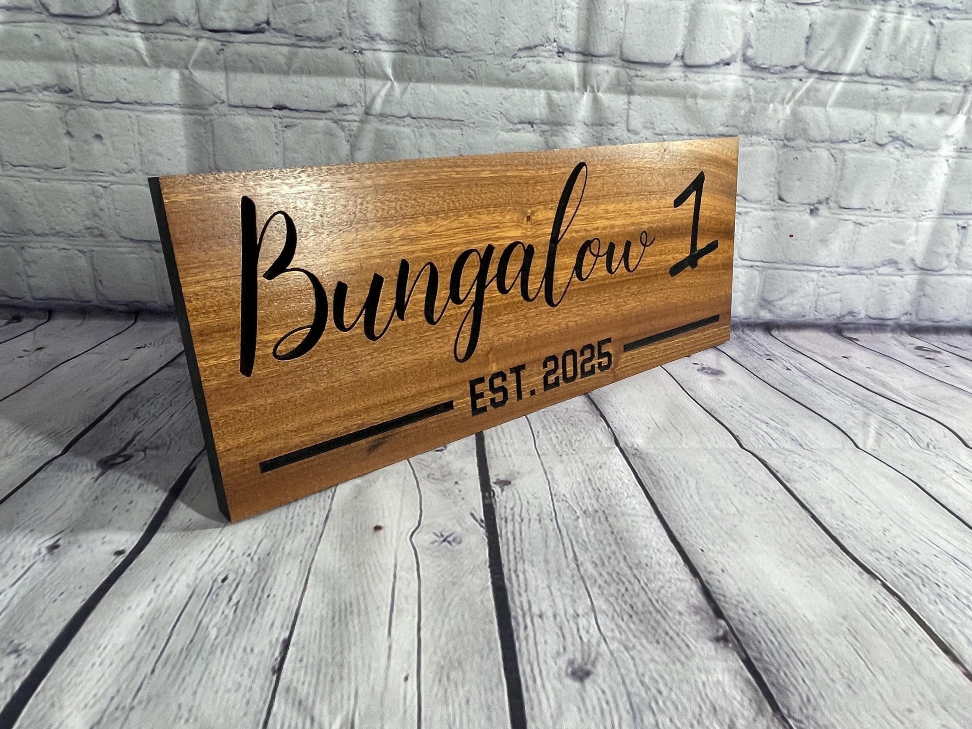 A wooden sign with the name "Bungalow 1" and the year "EST. 2005" printed on it, placed on a wooden surface against a brick wall background.