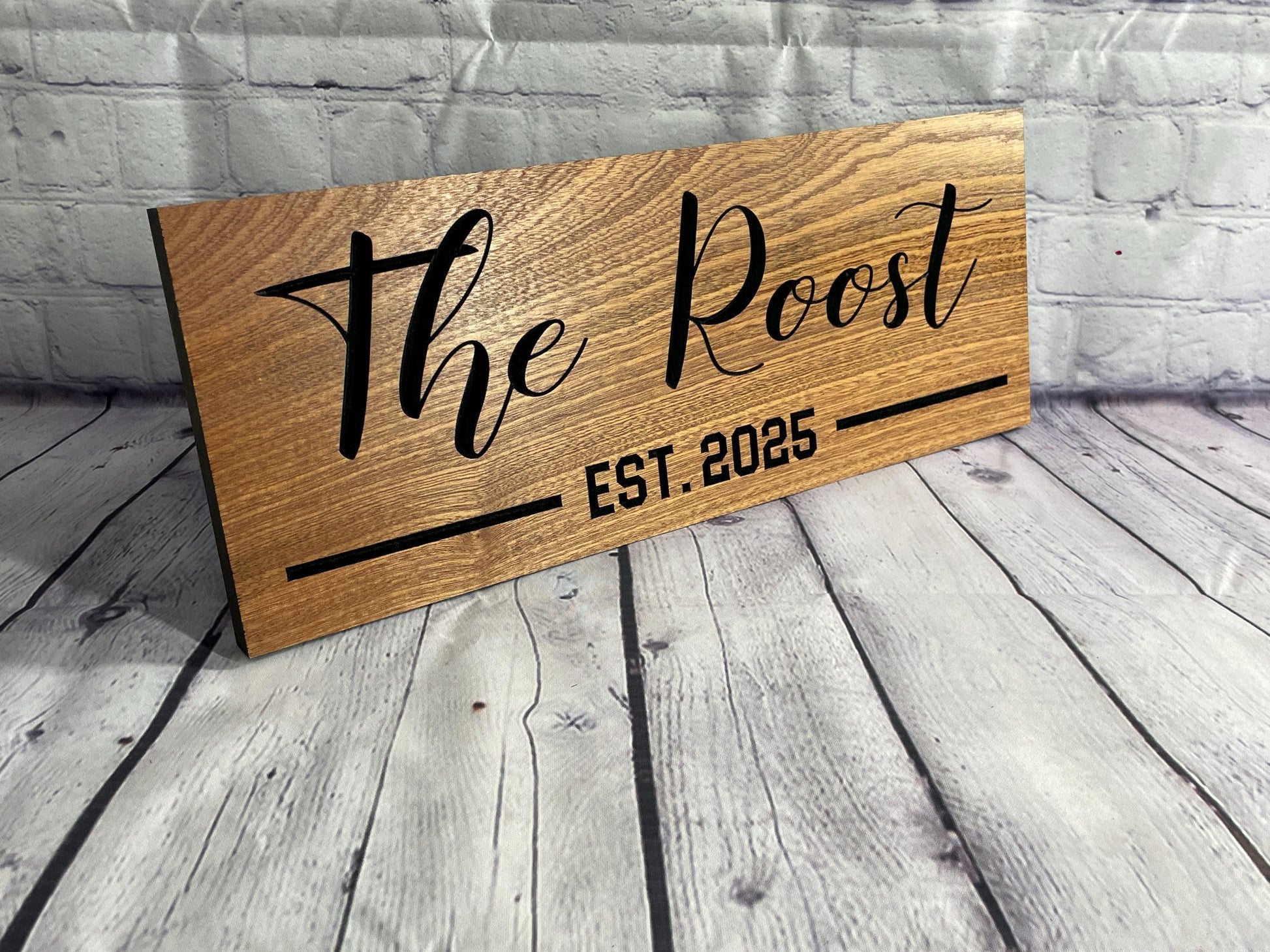A wooden sign with the name "The Roost" and the year "EST. 2025" printed on it, placed on a wooden surface against a brick wall background.