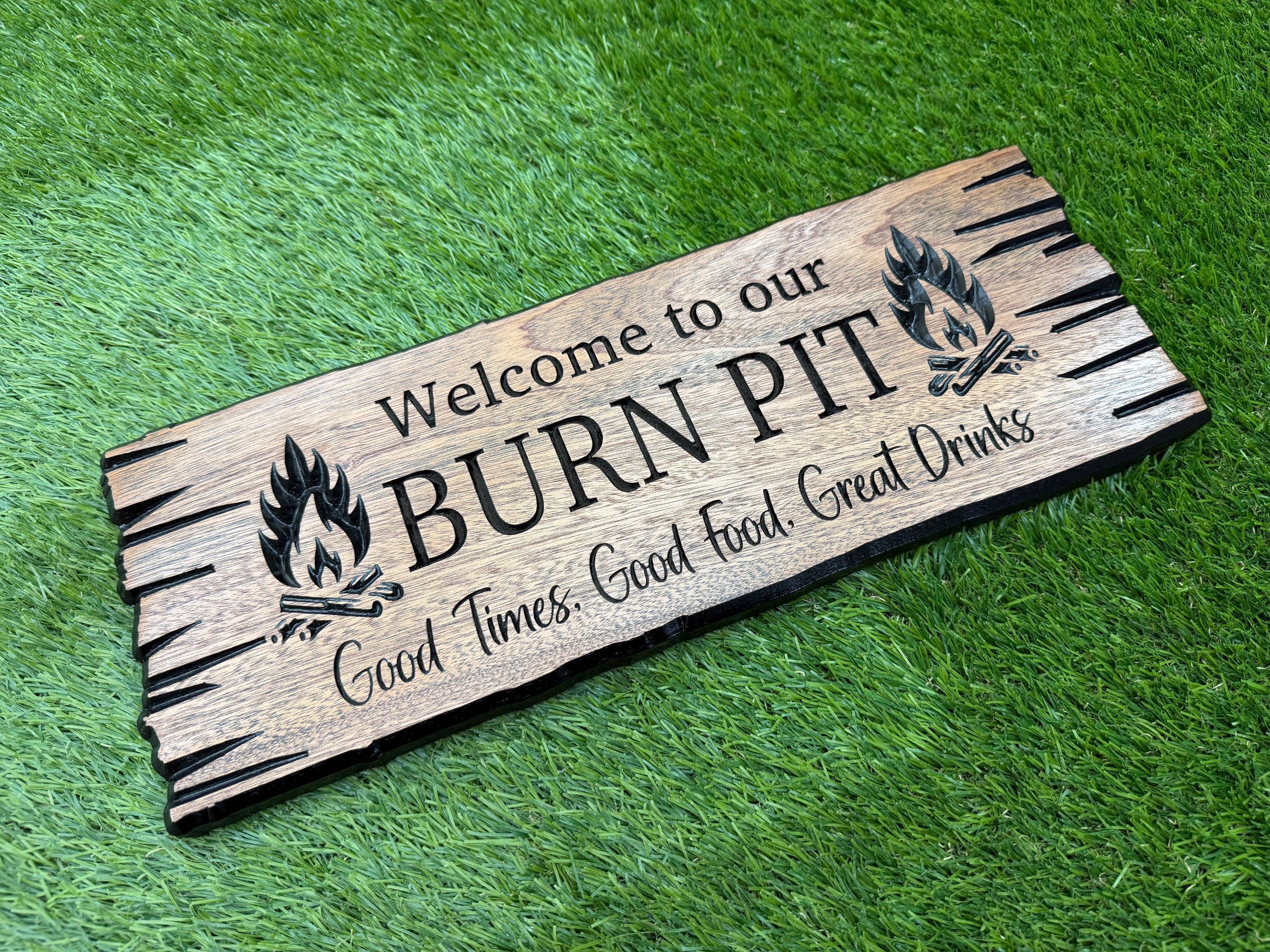 A wooden sign with the text "Welcome to our BURN PIT" and "Good times. Good food. Great drinks" on it, placed on a grassy surface.