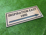 A wooden sign with the text "INSPIRATION EAST 1400" and an arrow pointing to the right, placed on a grassy surface.