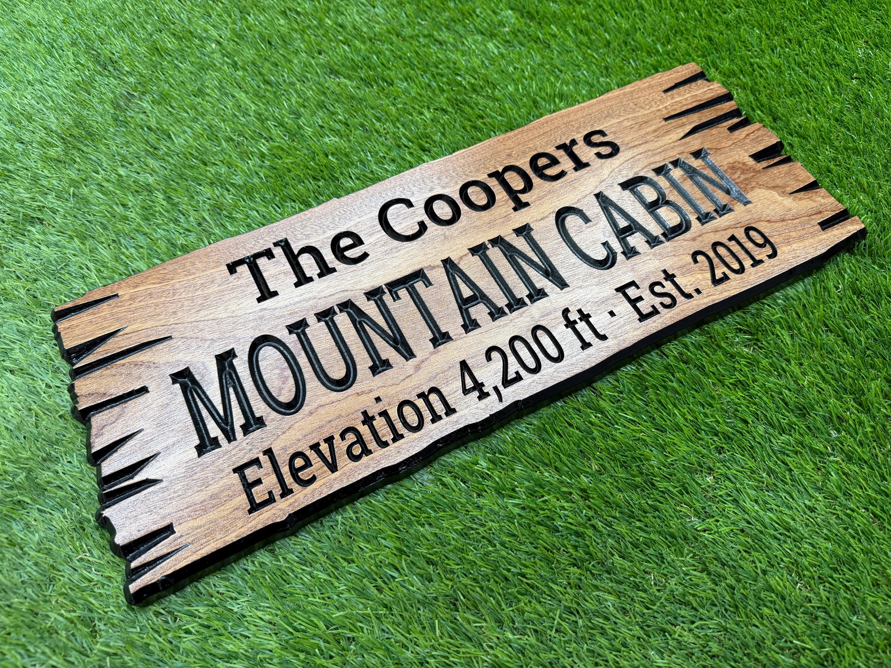 A wooden sign with the text "The Coopers Mountain Cabin Elevation 4,200 ft Est. 2019" engraved on it, placed on a grassy surface.