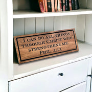 May include: A rectangular wooden sign with a dark brown wood grain finish and a black border. The text engraved in black reads: I CAN DO ALL THINGS THROUGH CHRIST WHO STRENGTHENS ME. PHIL. 4:13. The sign is positioned on a white shelf surface.
