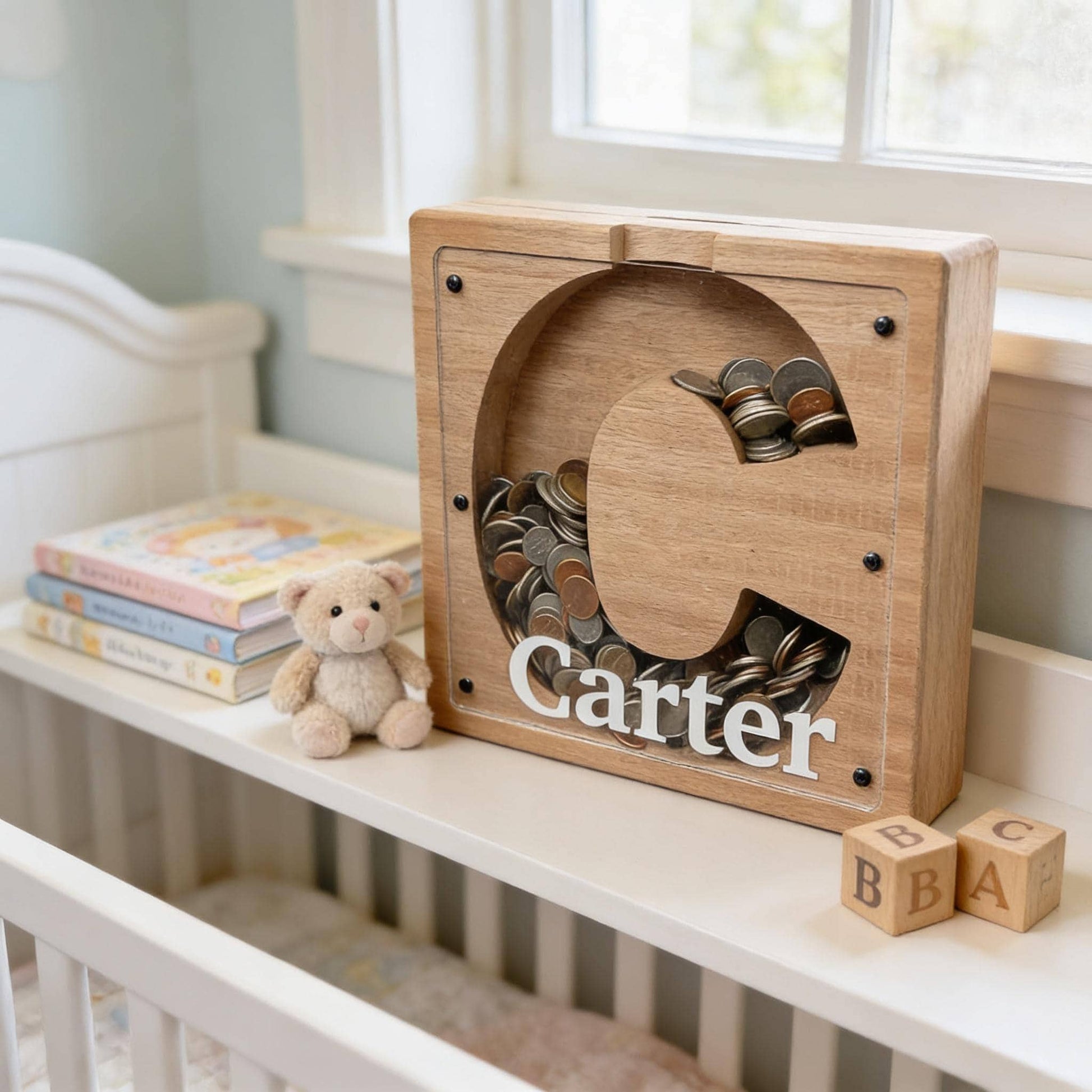 A wooden coin bank with the name " Carter" written on it, sitting on a white shelf next to a teddy bear and some blocks.