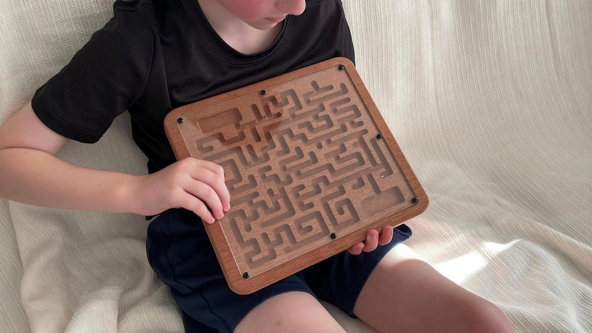 A young child is sitting on a white blanket, holding a wooden maze game in their hands.