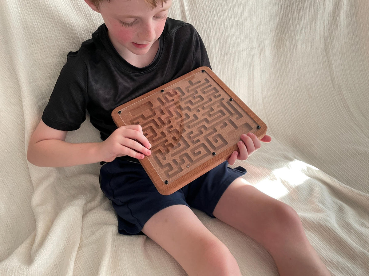 A young boy is sitting on a white blanket, holding a wooden maze puzzle in his hands.